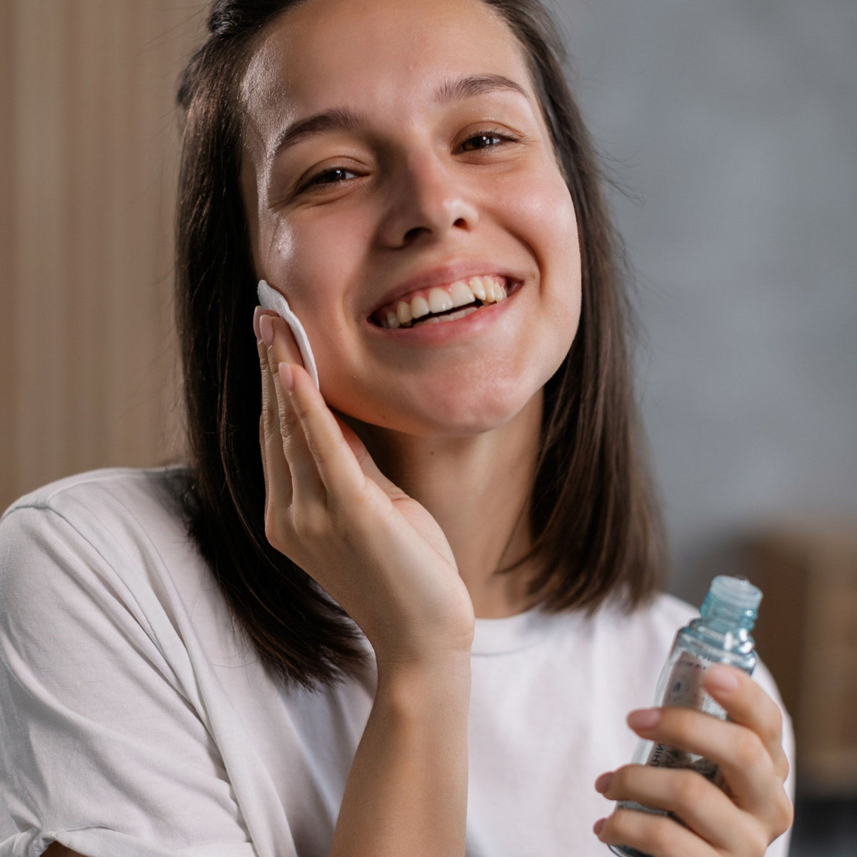 Ragazza sorridente che passa un dischetto di cotone con il tonico sulla guancia, con il flacone in mano